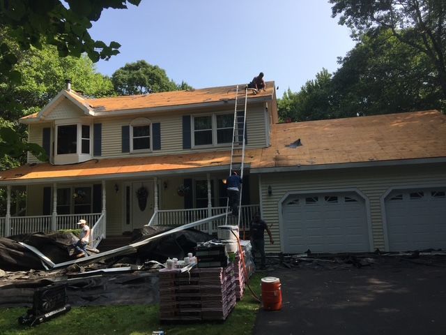 Roofers replacing shingles on a two-story house with a blue-gray facade; ladder and materials visible.