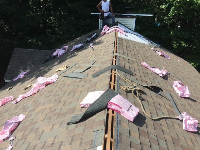 Roofer working on a brown shingle roof, insulation and supplies scattered.  Green trees in background.