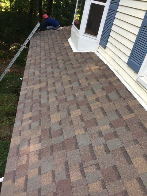 Roofer installing brown and tan asphalt shingles on a home's roof next to a bay window with blue shutters.