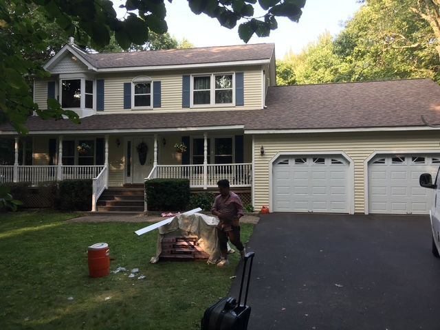 Two-story yellow house with white trim. Man working on lawn in front of garage. Dark roof and driveway.