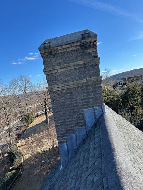A brick chimney, partially leaning, extends from a gray shingle roof against a blue sky.