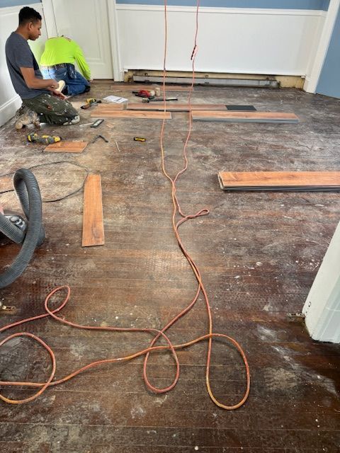 A person works on hardwood floor installation. Orange extension cord and tools are scattered on the wood floor.