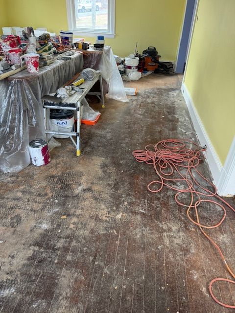 Room renovation in progress, dusty wooden floor, tools on table, extension cord, yellow walls, white trim.