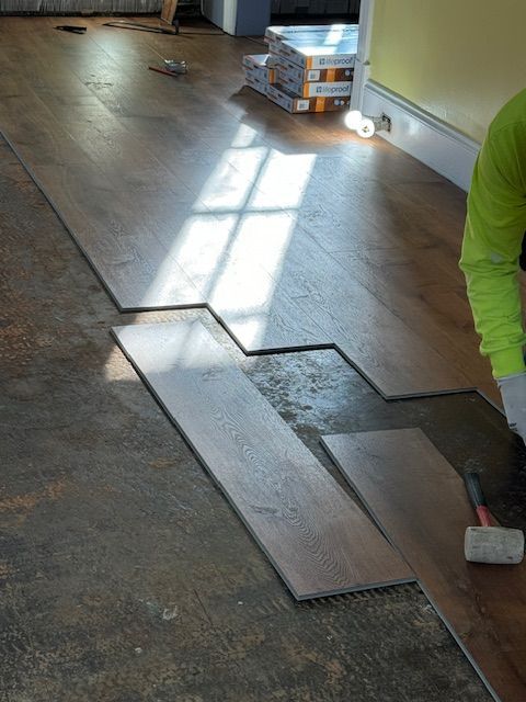 Installing wood-look flooring indoors. Person in neon green shirt lays planks on a patterned floor with sunlight.
