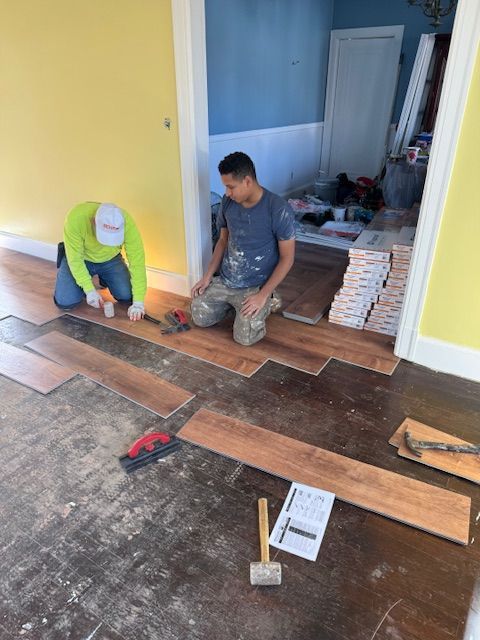 Two people installing wood flooring in a room. Tools and materials are scattered around.