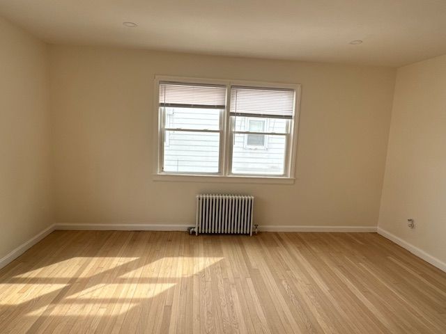 Empty room with window, radiator, and hardwood floor; beige walls.