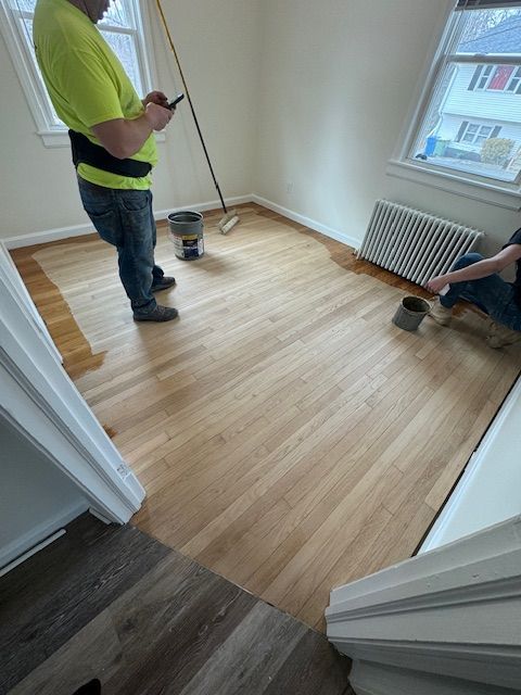 People staining a light wood floor in a room with windows and a radiator. One person is on their phone.