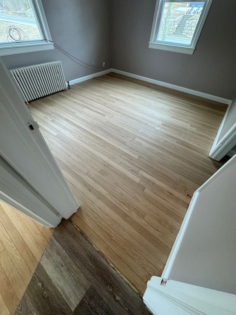 Empty room with hardwood floors, two windows, and pale pink walls. A radiator is visible.