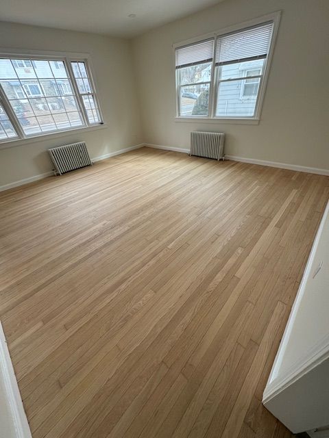 Empty room with wood floor, two windows, two radiators, and beige walls.