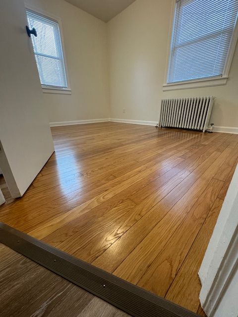 Hardwood-floored room with two windows, a radiator, and a partially open door. Beige walls, bright lighting.