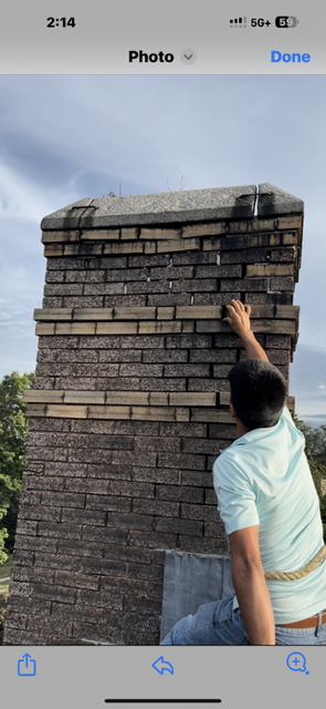 Person inspecting a brick chimney on a rooftop. The chimney is gray and beige, with a dark cap. The person wears a light blue shirt.