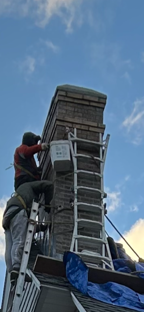 Two workers on a roof repairing a brick chimney. One is high on a ladder, working on the chimney.