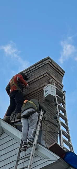 Two people working on a brick chimney from a ladder on a rooftop. Blue sky background.