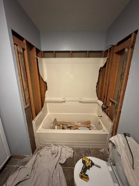 Bathroom undergoing renovation; drywall removed around the bathtub, exposing wooden studs. A power drill sits nearby.
