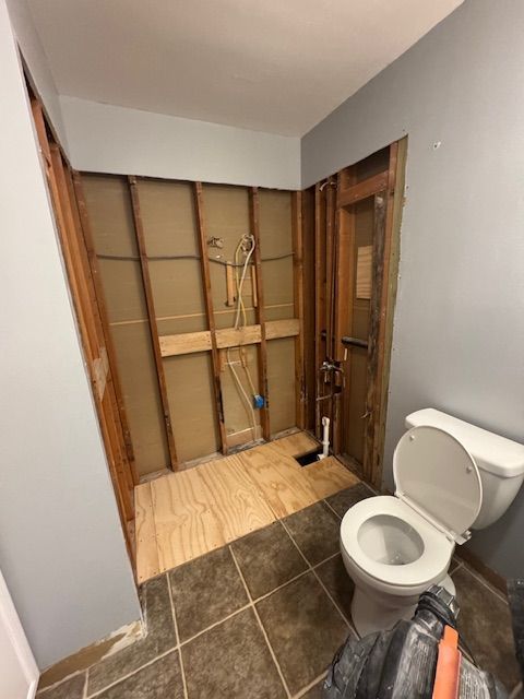 Bathroom renovation in progress. Walls open, exposed studs. Toilet visible. Gray and brown tile floor.