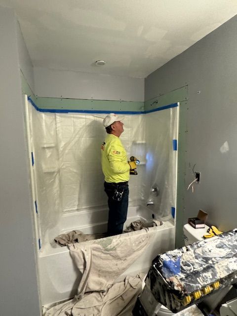 Man in a hard hat and yellow shirt working on a bathroom shower stall with a drill.