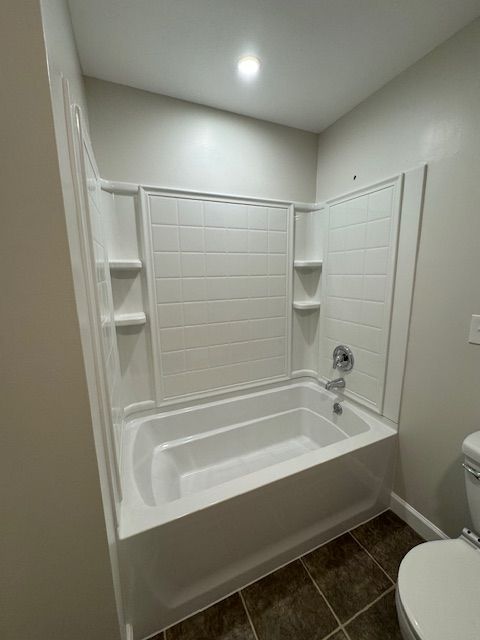 White tub and shower surround in a bathroom with dark floor tiles and a toilet.