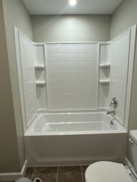 White bathtub and shower surround with built-in shelves in a bathroom, with a toilet visible.