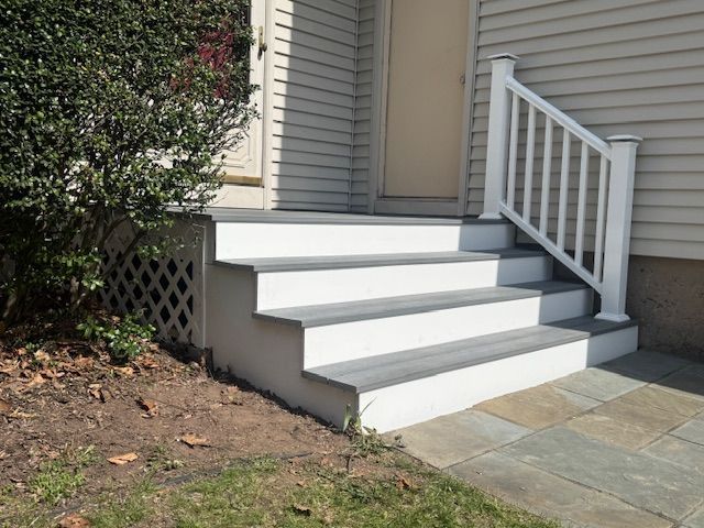 White steps with gray treads leading to a door, white railing on one side, greenery to the left, stone patio.