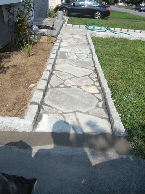 Stone walkway lined with cement borders leading to a house, with green grass to the right.
