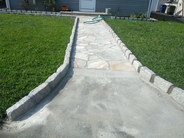 Concrete walkway with stone border and flagstone section, leading to a house entrance. Green lawn surrounds.