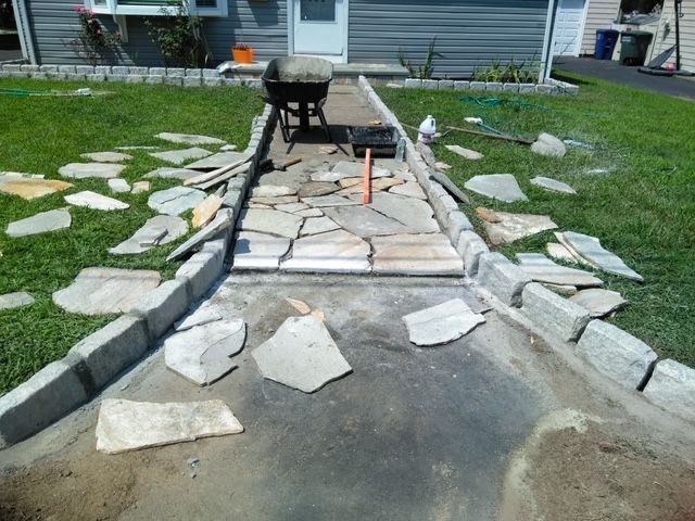Pathway construction: stone pavers laid on a concrete path with a wheelbarrow and tools, surrounded by green grass.