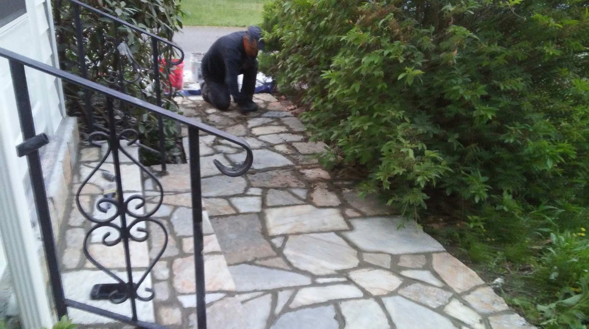 Person kneeling on a flagstone path, near a wrought-iron railing and green bushes.