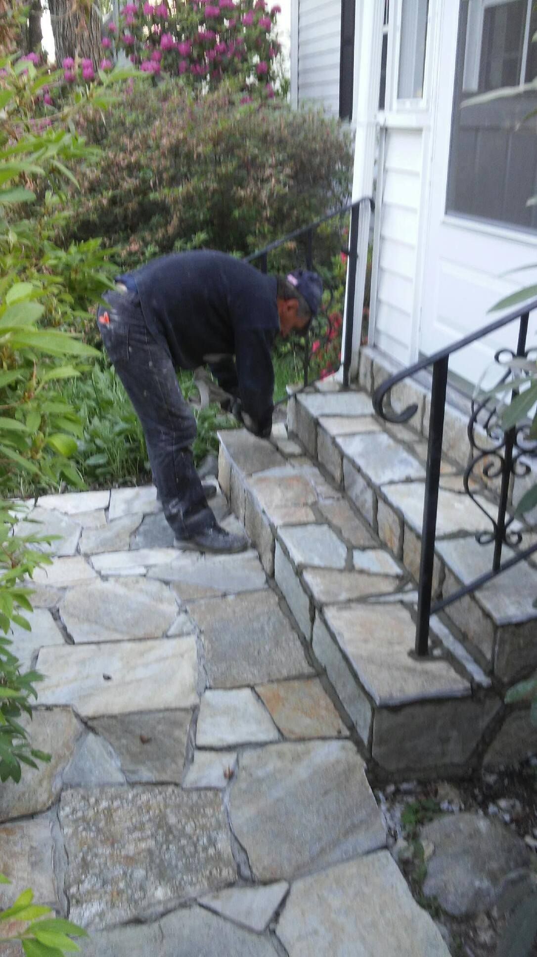 Man installing stone steps leading to a white house with a black railing, surrounded by greenery.