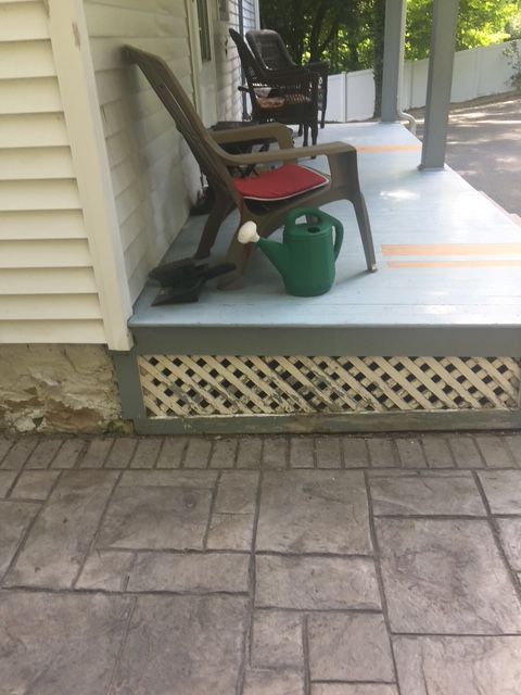 Porch with a chair, watering can, and lattice detail. Gray concrete pavers and white siding are visible.