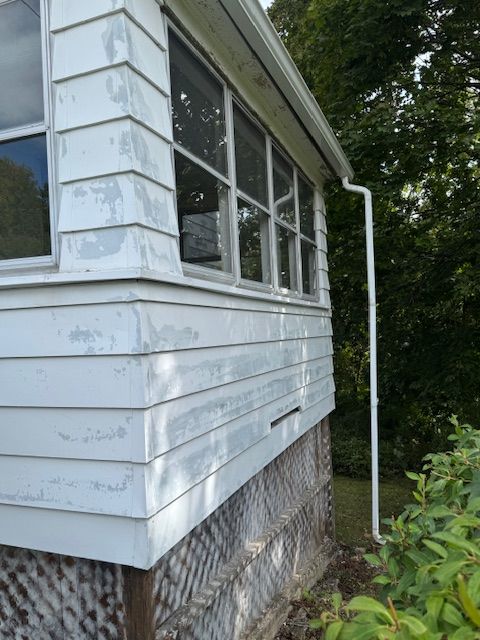 White house siding with peeling paint, windows, and a gutter, outdoors with foliage.