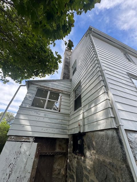 Side view of a weathered white house with a stone foundation and a tall chimney against a cloudy sky.