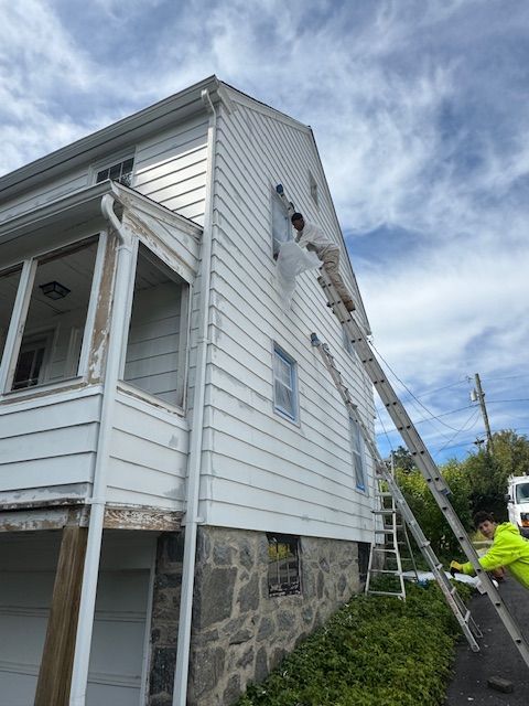 Person on ladder painting white siding of a two-story house with stone foundation, under a cloudy sky.