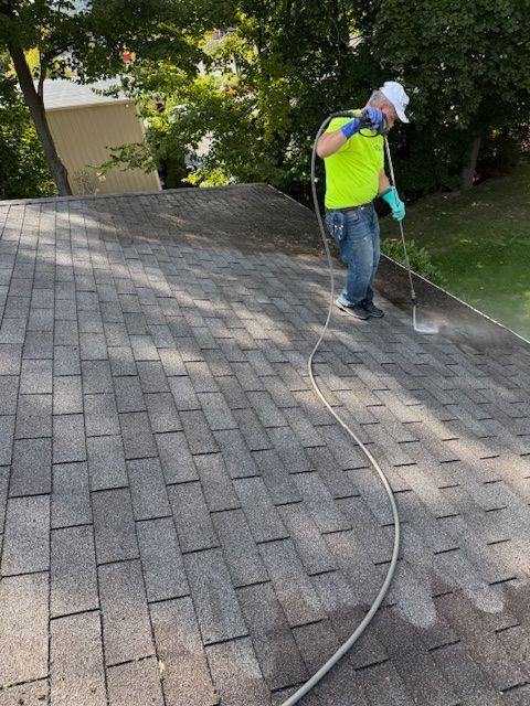 Person in safety gear power washing a gray shingle roof. Green shirt, blue jeans, white hat, and a hose.