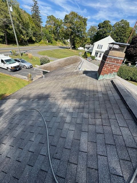 A roof covered in gray asphalt shingles, with moss, overlooking a street and houses under a blue sky.