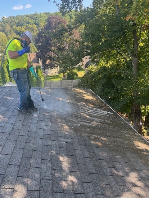 Man in safety vest power washing a shingled roof on a sunny day.
