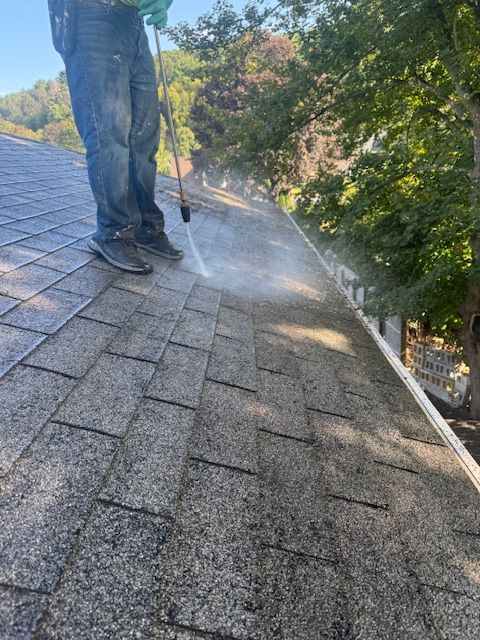Person pressure washing a gray shingle roof. Water sprays across the shingles.