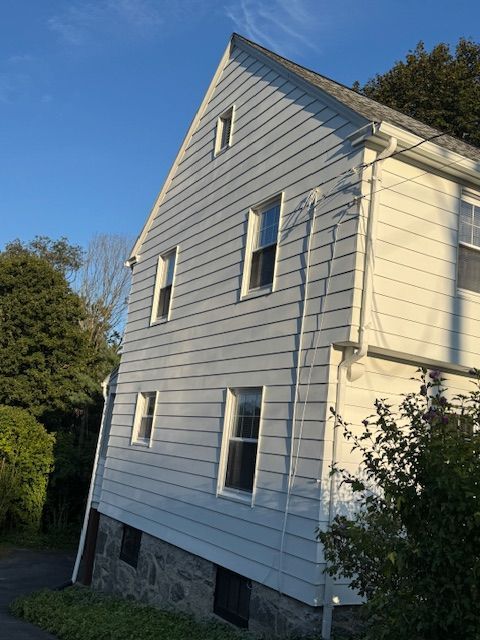 Side view of a two-story house with gray siding, white trim, and a stone foundation, under a blue sky.