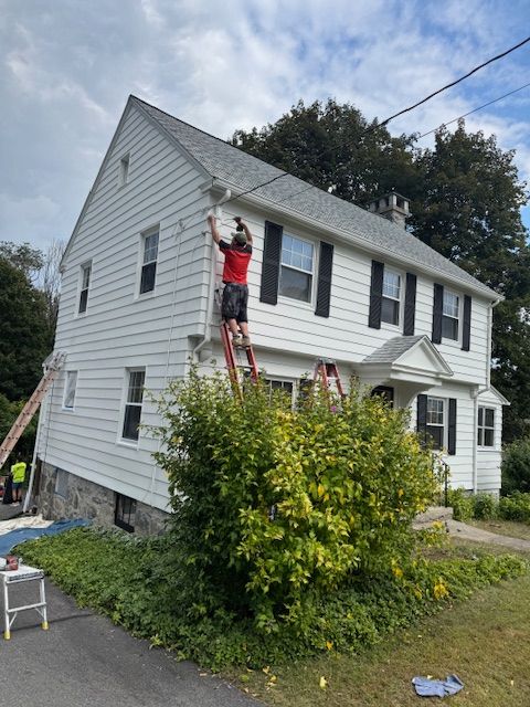 Person on a ladder cleaning a white house's gutters, sunny day.