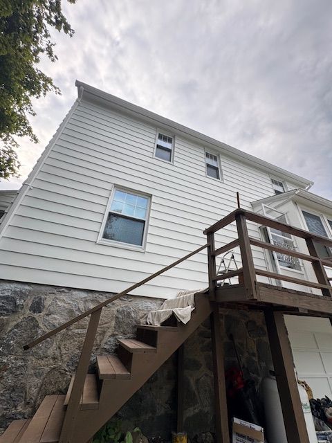 White house with white siding, stone foundation, and wooden stairs leading up to a deck. Cloudy sky.