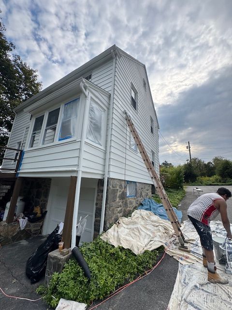 House being painted; painter on a tarp, ladder leaning against the white siding. Blue sky, bushes in front.