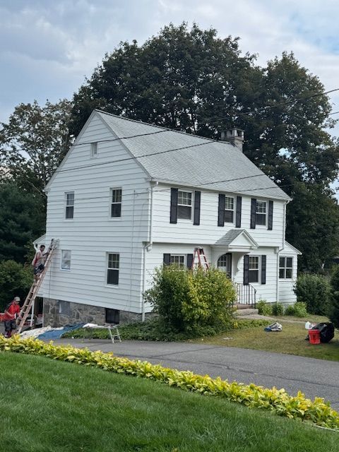 White house with black shutters, roofers on a ladder, dark green trees.