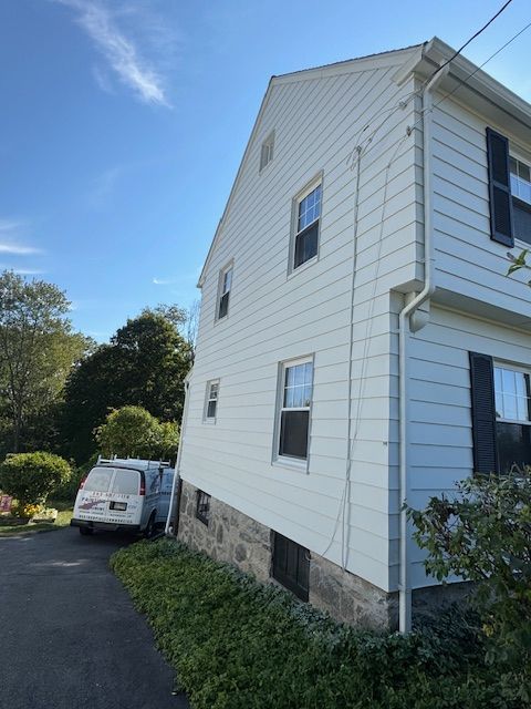 White house exterior with three windows, a van, and a stone foundation on a sunny day.