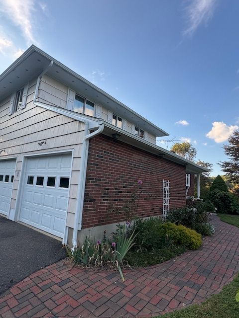 Two-story house with white garage doors and brick siding. Red brick walkway curves along the right side.