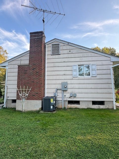 Side view of a house with a brick chimney, air conditioning unit, and antenna. White siding and green grass.