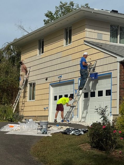 Three people painting the exterior of a two-story house. One on a ladder, two on ladders leaning against the wall.