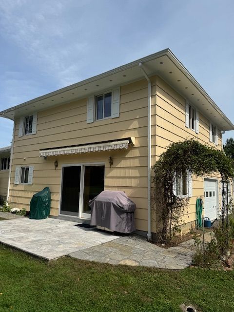 Two-story yellow house with a sliding door, patio, and garage; a covered grill and a green container are visible.