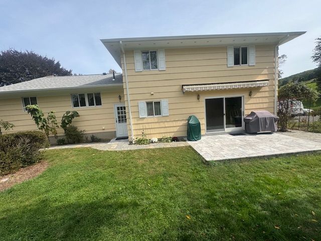 Back of a two-story yellow house with a patio, lawn, and covered grill under a blue sky.