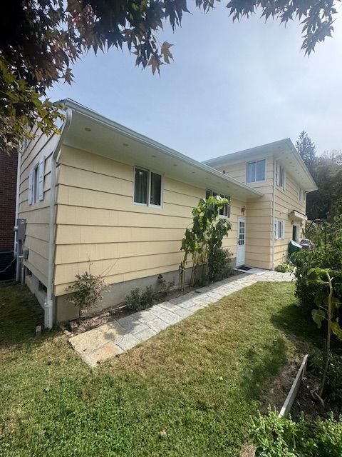 Yellow-sided two-story house with white trim, a walkway, and green grass and foliage under a blue sky.