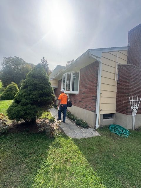 Person in orange shirt walking toward a brick house with a curved window and green hose on the grass.