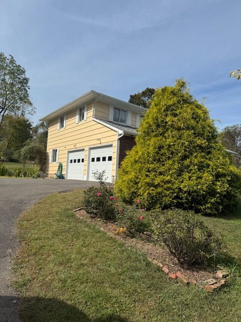 Yellow two-story house with two-car garage, driveway, and large green bush, set on a grassy lawn with roses.